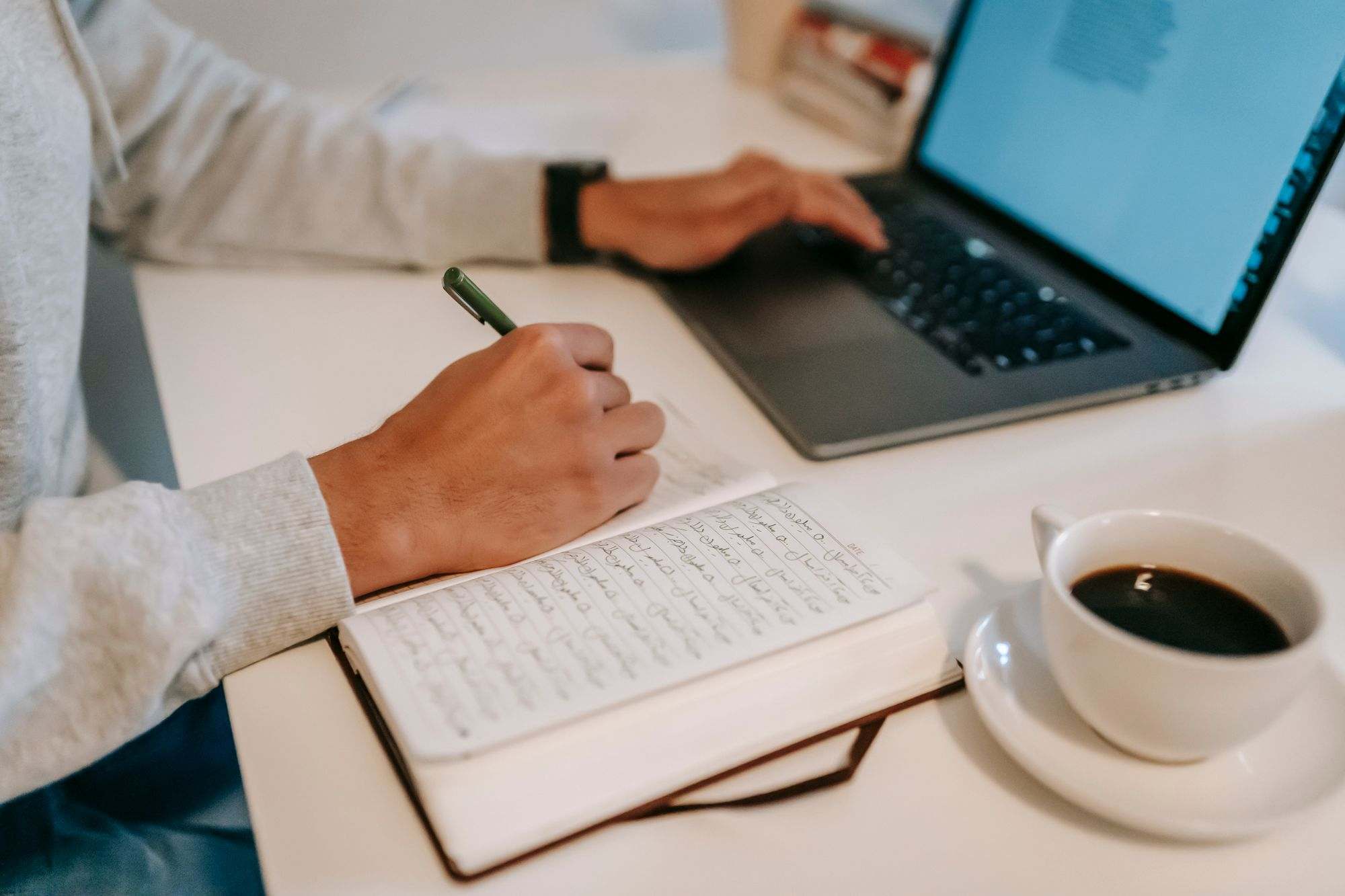 Hands writing notes beside a laptop and a cup of coffee