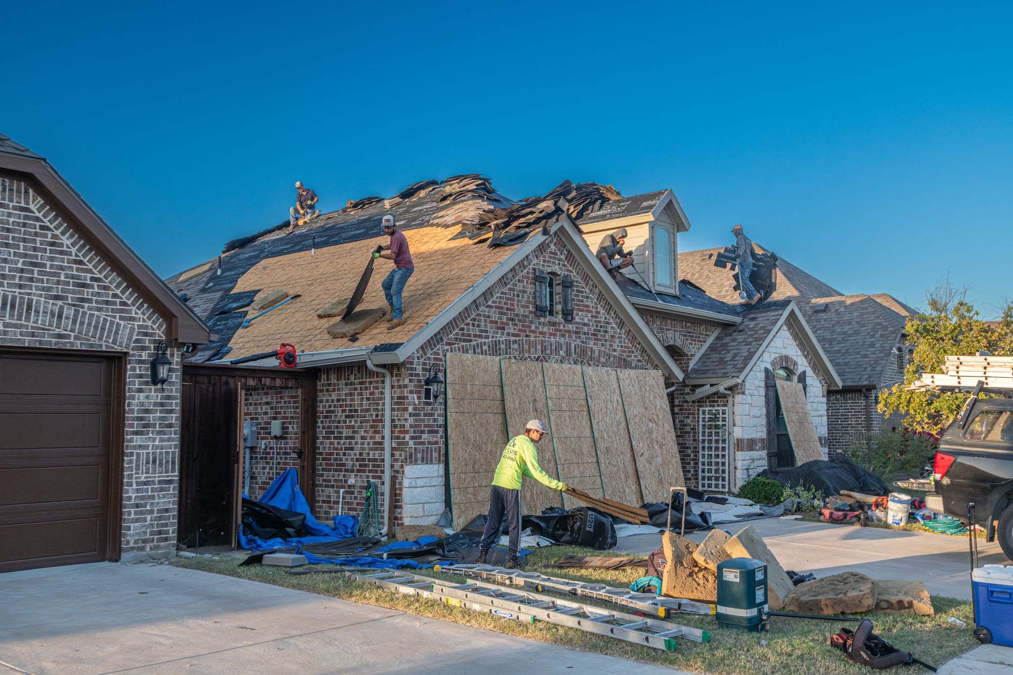 A crew replacing the roof of a brick house, with materials stacked in the driveway